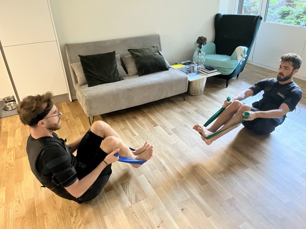 Deux hommes en séance d’électrostimulation (EMS) à domicile à Genève, réalisant un exercice de renforcement musculaire avec élastiques de résistance